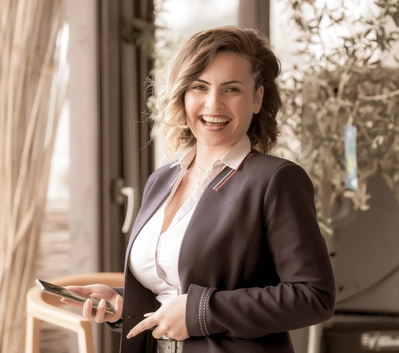 Alexa Nica râzând, în sacou bleumarin, lângă o fereastră cu un măslin, lumină caldă